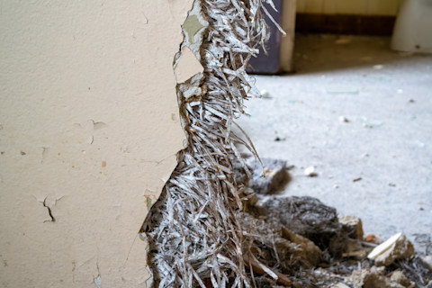 A broken interior wall of a house exposes old, stringy asbestos. The floor behind it is covered in rubble and stones, suggesting ongoing refurbishment work.