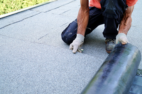 A worker kneels on a flat roof, unrolling a sheet of roofing material. They wear gloves and a red shirt. The background shows green foliage.